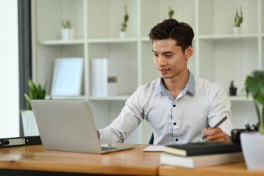 Dizüstü bilgisayardaki finansal verileri analiz eden Asyalı erkek yatırımcıya konsantre ol ve not defterine not al.