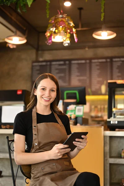 Image of successful small business owner using digital tablet and looking at camera while sitting in her modern coffee shop.