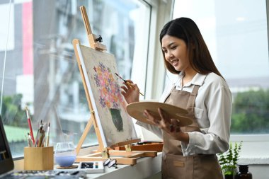 Resim stüdyosunda palet ve fırçayla resim yapan önlüklü hoş bir kadın sanatçı. Eğitim, hobi, sanat konsepti.