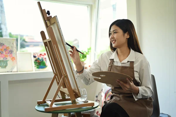 Pleasant female artist in apron with palette and brush painting on canvas at art studio. Education, hobby, art concept.