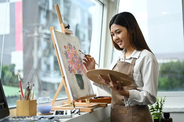 Pleasant female artist in apron with palette and brush painting on canvas at art studio. Education, hobby, art concept.