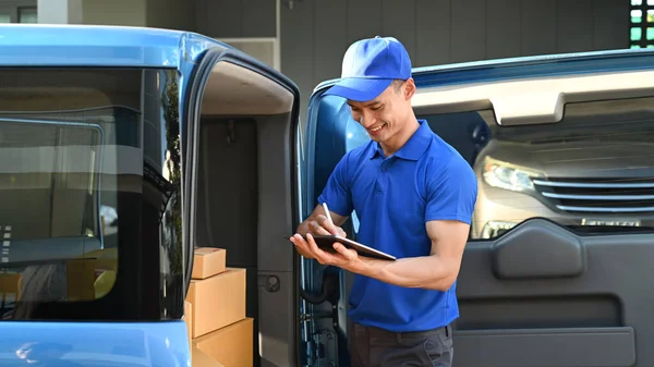 Delivery man in blue uniform using digital tablet next to open delivery van. Shipping and delivery service concept.
