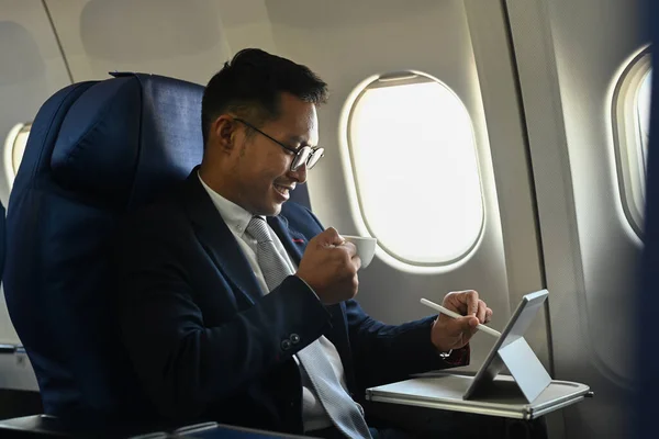 Smiling businessman drinking coffee and using using tablet and during business travel flight.