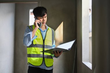 Yansıtıcı yelek giyen mühendis inşaat alanında duruyor ve cep telefonuyla konuşuyor..
