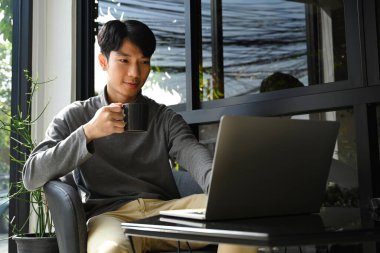 Yakışıklı Asyalı adam sıcak kahve içiyor ve laptopta haber ve e-postaları kontrol ediyor..