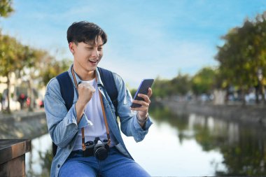 Asyalı gezgin güzel bir günde şehir parkında otururken akıllı telefon kullanıyor. Seyahat, tatil ve seyahat kavramı.