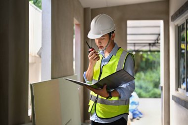 Civil engineer communicating with walkie talkie and working with blueprint at construction site.