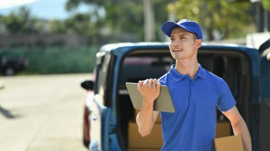 Portrait of delivery man wearing blue uniform with cardboard in hands and using digital tablet.