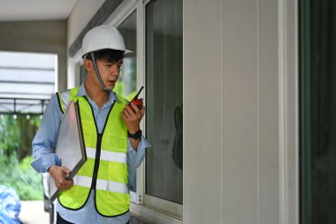 Engineer in safety helmet communicating with walkie talkie and checking building construction progress.