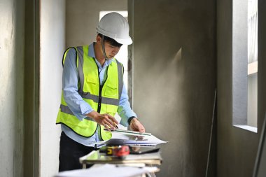 Engineer man in safety helmet working at blueprints and checking building construction progress at construction site.