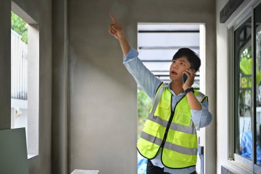 Asian male civil engineer manager having phone conversation and inspecting building construction site.