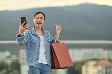 Cheerful woman making video call on mobile phone, standing on terrace with beautiful evening sky on background.