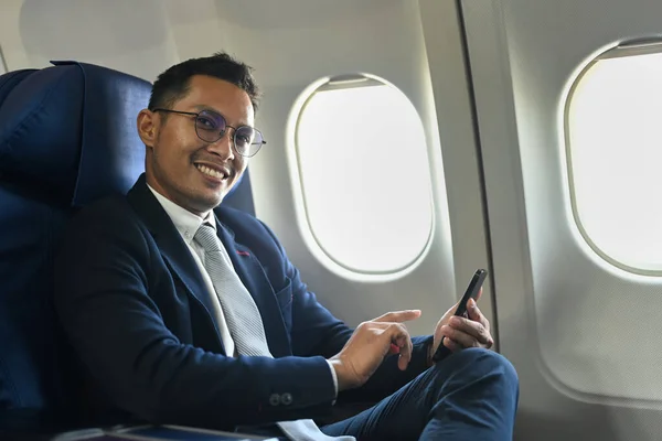Happy millennial businessman sitting in the airplane next to the window and using smart phone.