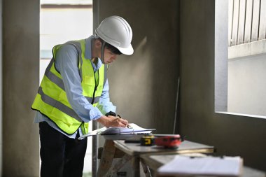 Engineers man wearing safety helmet and vest working with blueprints in building construction site.