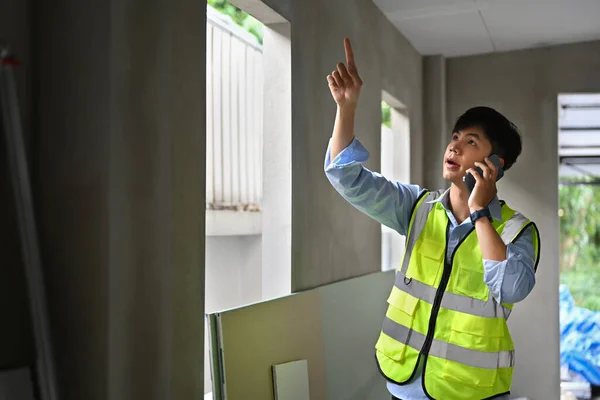 Asian male engineer supervisor having phone conversation while inspecting building construction site.