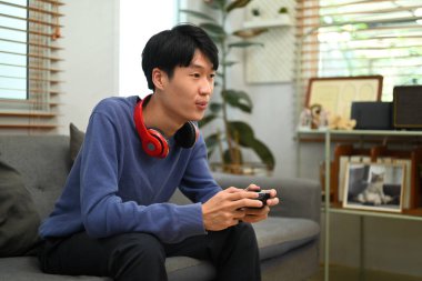 Happy young asian man player holding gamepad controller playing video game in living room.