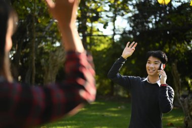 Friendly asian man walking in public park with mobile phone and greeting friend with hand raised up.