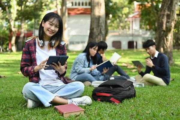 Cheerful asian student woman using digital tablet on green meadow in campus. Education, youth and community concept.