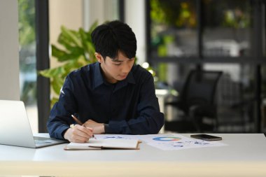 Focused asian male economist using laptop and checking statistic document at office desk.