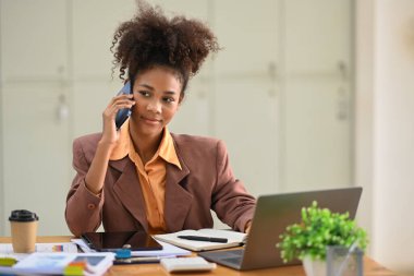 Smiling African American female entrepreneur working on laptop computer and having conversation on mobile phone.