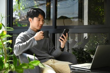 Pleasant young asian man sitting outdoor coffee shop and chatting online in social network on smart phone.