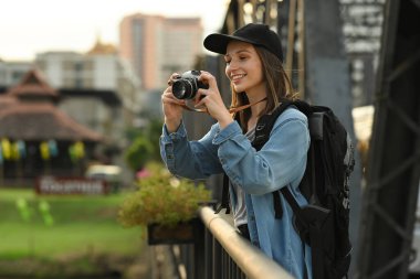 Cheerful female travel blogger recording video vlog with camera while standing on bridge with scenery view of river in Thailand.