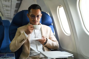 Businessman in elegant suit drinking coffee, relaxing in comfortable seat next to aircraft cabin window.