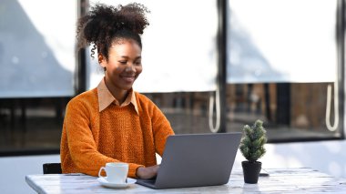 Cheerful African American female freelancer sitting outside cafe and checking email, working online on her laptop.