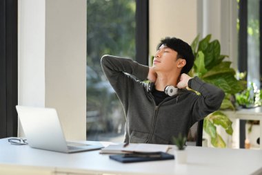 Relaxed young asian man worker sitting back in his chair with closed eyes and hands clasped behind head.