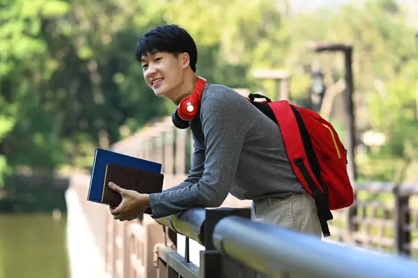 Relaxed Asian Man College Student Backpack Headphone Standing Bridge River — Zdjęcie stockowe