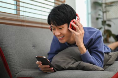 Happy young asian man listening to music in headphone and using smart phone on comfortable couch.