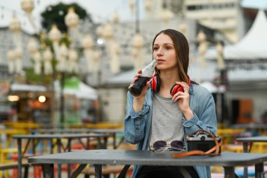 Beautiful woman in jean jacket drinking cola from glass bottle while sitting at outdoor street food restaurant.