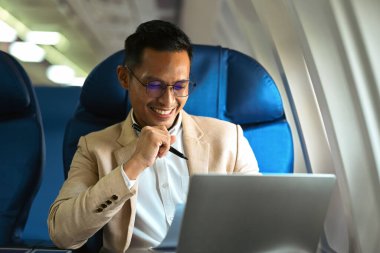 Image of smiling businessman in elegant luxury suit working with laptop in aircraft during business travel.
