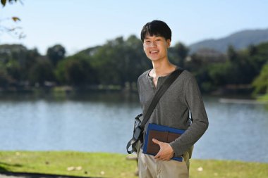 Portrait of asian student man standing near lake with mountains sunny landscape on background. People and recreation in nature.