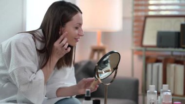 Attractive caucasian woman wearing bathrobe applying skincare cream on face and looking in mirror. Self care and daily beauty routine.