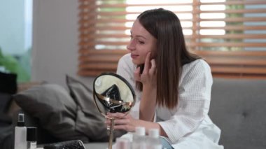 Charming caucasian woman wearing bathrobe applying nourishing cream on her face and enjoying facial skincare routine.