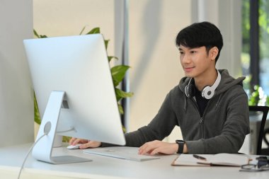 Young creative man wearing gray hoodie looking at computer monitor, working remotely from home.