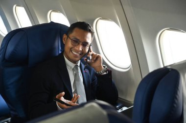 Confident businessman in black suit sitting comfortable seat in airplane cabin and talking on mobile phone.