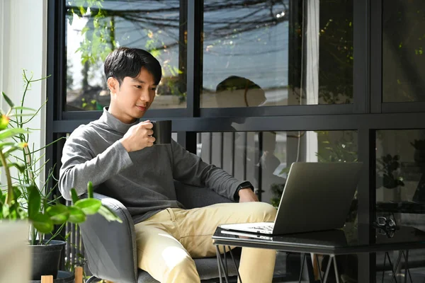 Relaxed young asian man sitting outdoor coffee shop and reading email or online news on laptop.