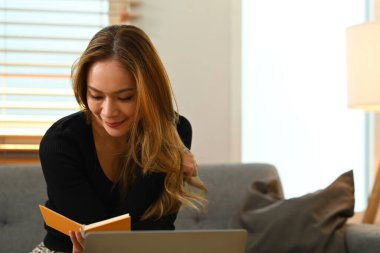 Relaxed young woman sitting in comfortable couch at home and reading interesting book, spending leisure weekend at home.