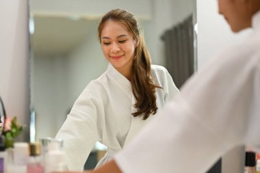 Smiling millennial woman in bathrobe standing front of mirror and making daily beauty routine at home.