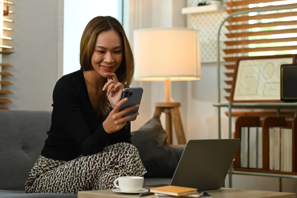Satisfied woman using smart phone chatting with friends in social network while relaxing on couch at home.