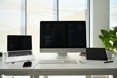 Computer monitors with programming code on white office desk at software development company.