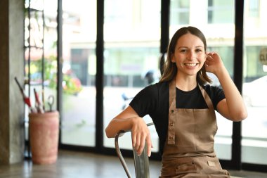 Confident caucasian female entrepreneur in apron sitting in her coffee shop and smiling to camera.