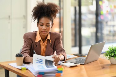 Concentrated African American female economist analyzing financial data and using laptop computer at work station.