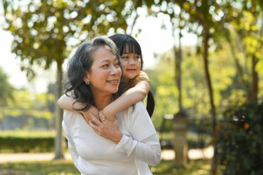 Smiling mature grandmother giving piggyback ride to cute little granddaughter during walking on in the park.