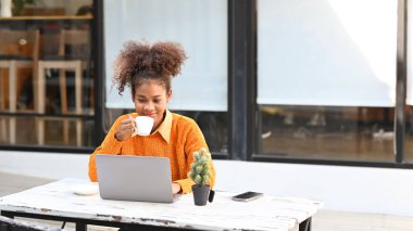 Smiling young African woman relaxing in outdoor cafe and using laptop on white wooden table.