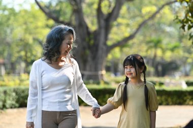 Smiling grandmother and her little grandchild walking together surrounded by trees during sunny day.
