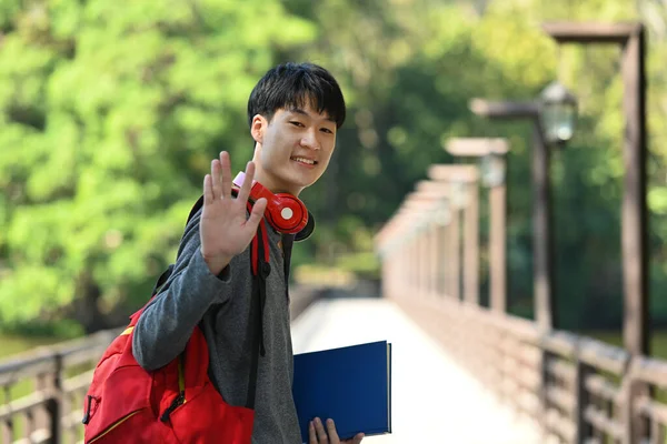 Photo Smiling Asian Man College Student Backpack Books Waving Hand — Zdjęcie stockowe