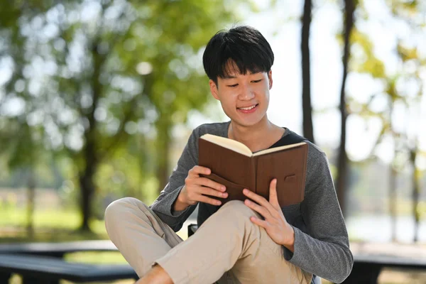 Happy Young Asian Man Reading Interesting Book While Sitting Bench — Zdjęcie stockowe